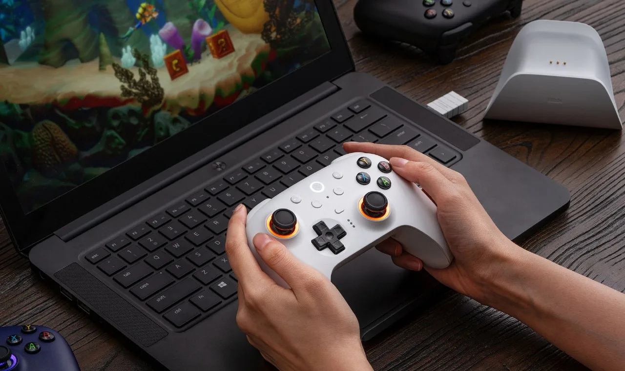 White 8BitDo Ultimate 2 controller in hands next to a laptop, the dongle and a charging dock