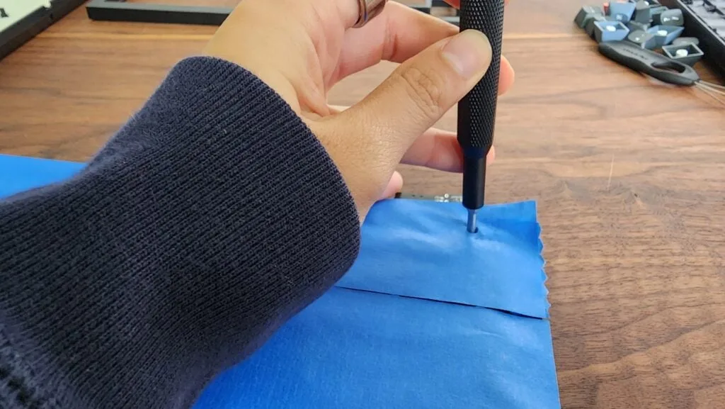 Screwdriver poking through painter's tape to open a screw hole on a taped keyboard PCB